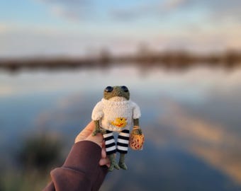 May include: A knitted frog toy wearing a white sweater with a yellow and orange design and black and white striped pants. The frog is holding a small brown bag.