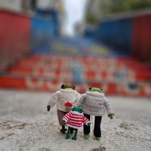 May include: Three knitted frog figurines, two wearing sweaters and one wearing a striped shirt, standing on a concrete surface.