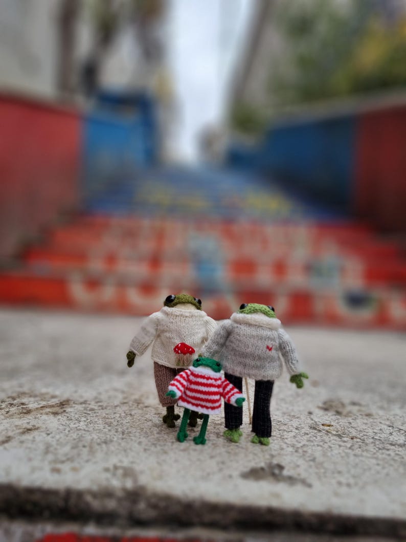 May include: Three knitted frog figurines, two wearing sweaters and one wearing a striped shirt, standing on a concrete surface.