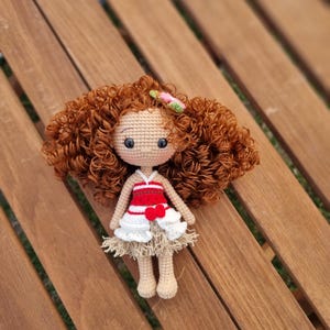 May include: A crocheted doll with brown curly hair, wearing a red and white dress with a flower in her hair. The doll is sitting on a wooden surface.