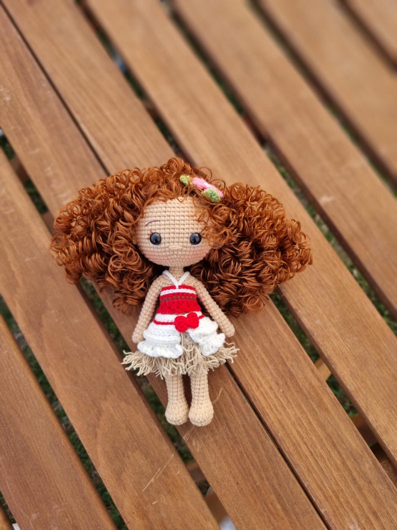 May include: A crocheted doll with brown curly hair, wearing a red and white dress with a flower in her hair. The doll is sitting on a wooden surface.