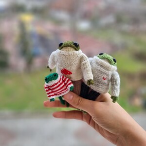 May include: Three knitted frog plushies wearing sweaters. The largest frog is wearing a white sweater with a red heart, the middle frog is wearing a white sweater with a red heart, and the smallest frog is wearing a red and white striped sweater.