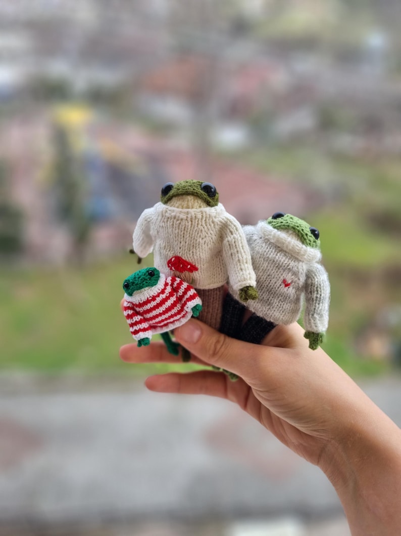 May include: Three knitted frog plushies wearing sweaters. The largest frog is wearing a white sweater with a red heart, the middle frog is wearing a white sweater with a red heart, and the smallest frog is wearing a red and white striped sweater.