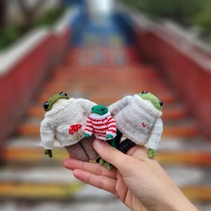 Op de afbeelding: Drie gebreide kikker knuffels die truien dragen. Eén kikker draagt een witte trui met een rode paddenstoel, een andere draagt een rood-wit gestreepte trui en de derde draagt een witte trui met een rood hart.