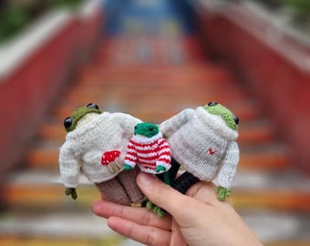 May include: Three knitted frog plushies wearing sweaters. One frog is wearing a white sweater with a red mushroom, another is wearing a red and white striped sweater, and the third is wearing a white sweater with a red heart.