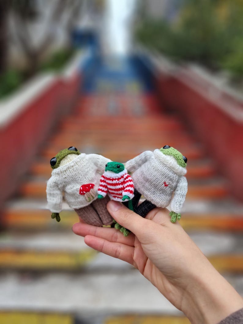 May include: Three knitted frog plushies wearing sweaters. One frog is wearing a white sweater with a red mushroom, another is wearing a red and white striped sweater, and the third is wearing a white sweater with a red heart.