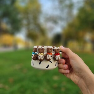 May include: A white ceramic mug with black markings, featuring four small crocheted monkey figures hanging from the rim. The monkeys are wearing colorful hats and have brown fur.