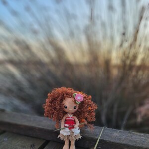 May include: A crocheted doll with brown curly hair, wearing a red and white top and a white skirt, holding a stick. The doll is sitting on a wooden surface with a blurred background of tall grass.