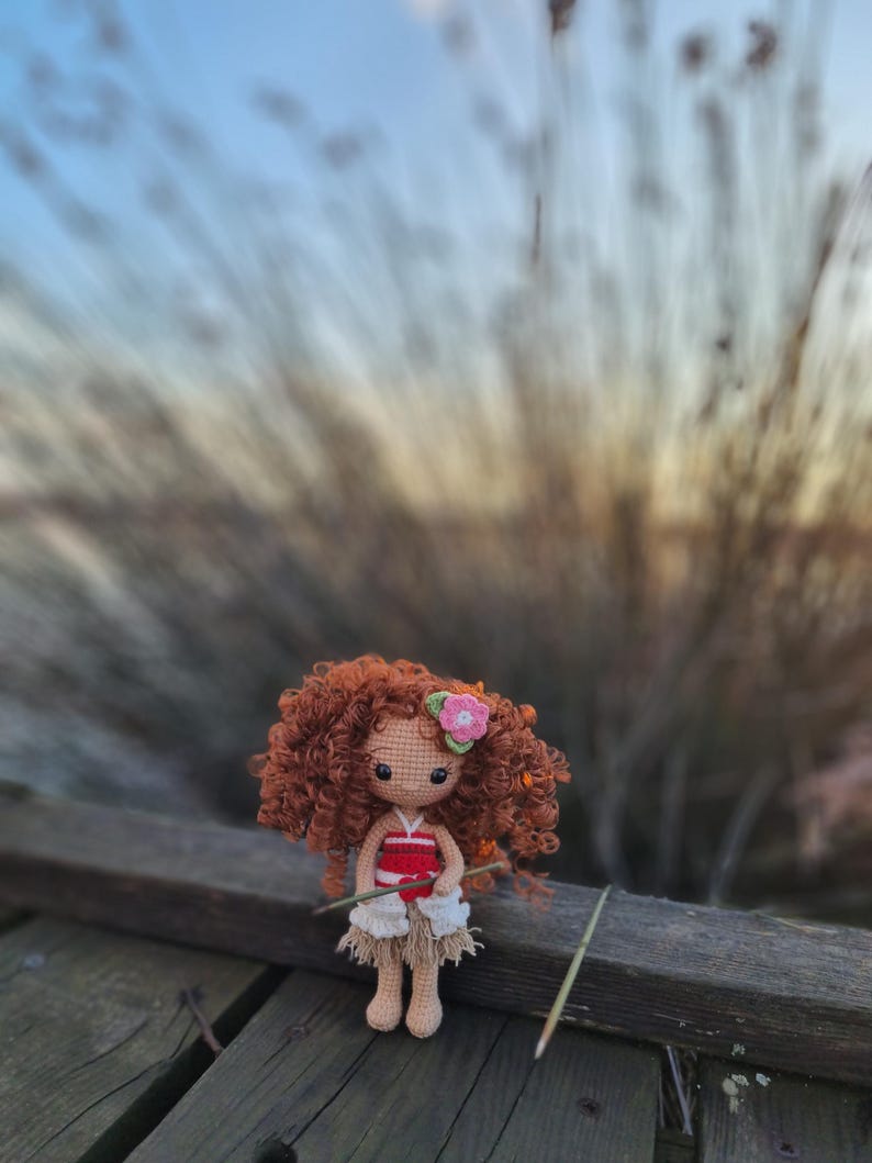 May include: A crocheted doll with brown curly hair, wearing a red and white top and a white skirt, holding a stick. The doll is sitting on a wooden surface with a blurred background of tall grass.