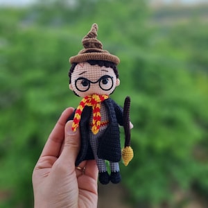 May include: A crocheted amigurumi doll of a wizard wearing a brown pointed hat, black robes, a red and yellow scarf, and round glasses. The doll is holding a brown stick with a yellow star on the end.