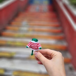 May include: A small green knitted frog wearing a red and white striped sweater. The frog is being held in a hand.