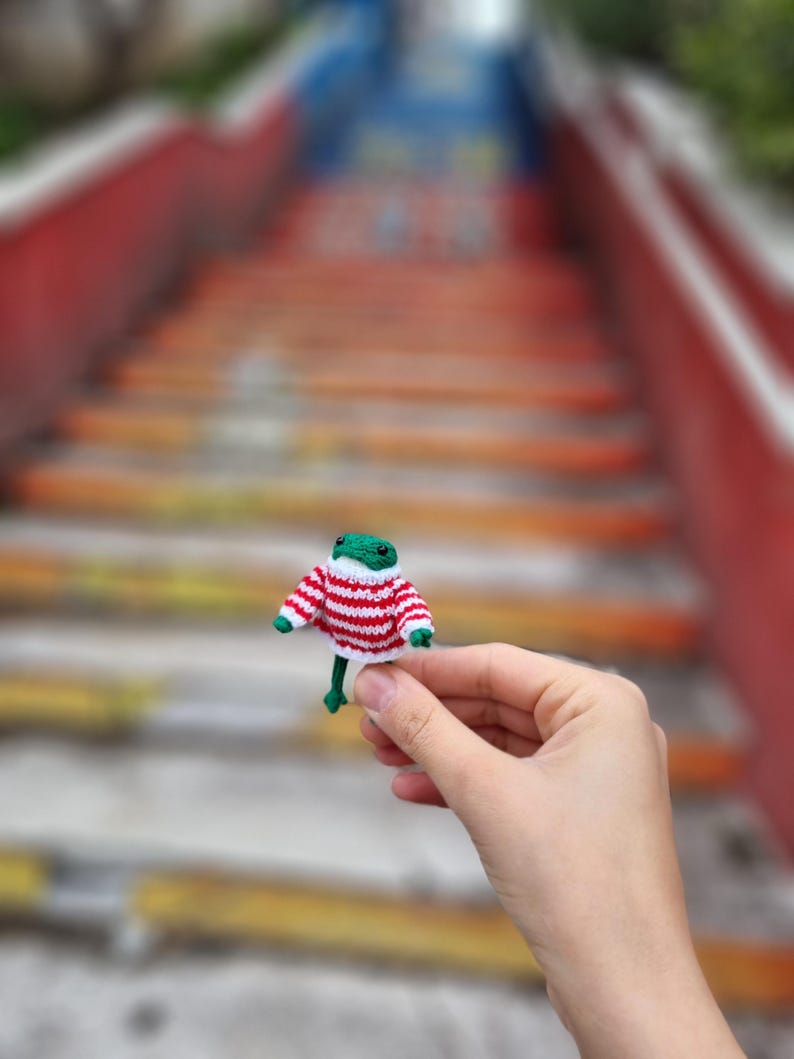 May include: A small green knitted frog wearing a red and white striped sweater. The frog is being held in a hand.