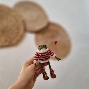 May include: A handmade green frog figurine wearing a red and white striped sweater and brown boots. The frog has a white collar and is holding a small red object. The frog is a whimsical, handcrafted item.