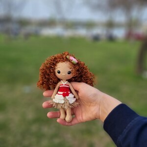 May include: A crocheted doll with brown curly hair, wearing a red and white dress with a flower on her head.