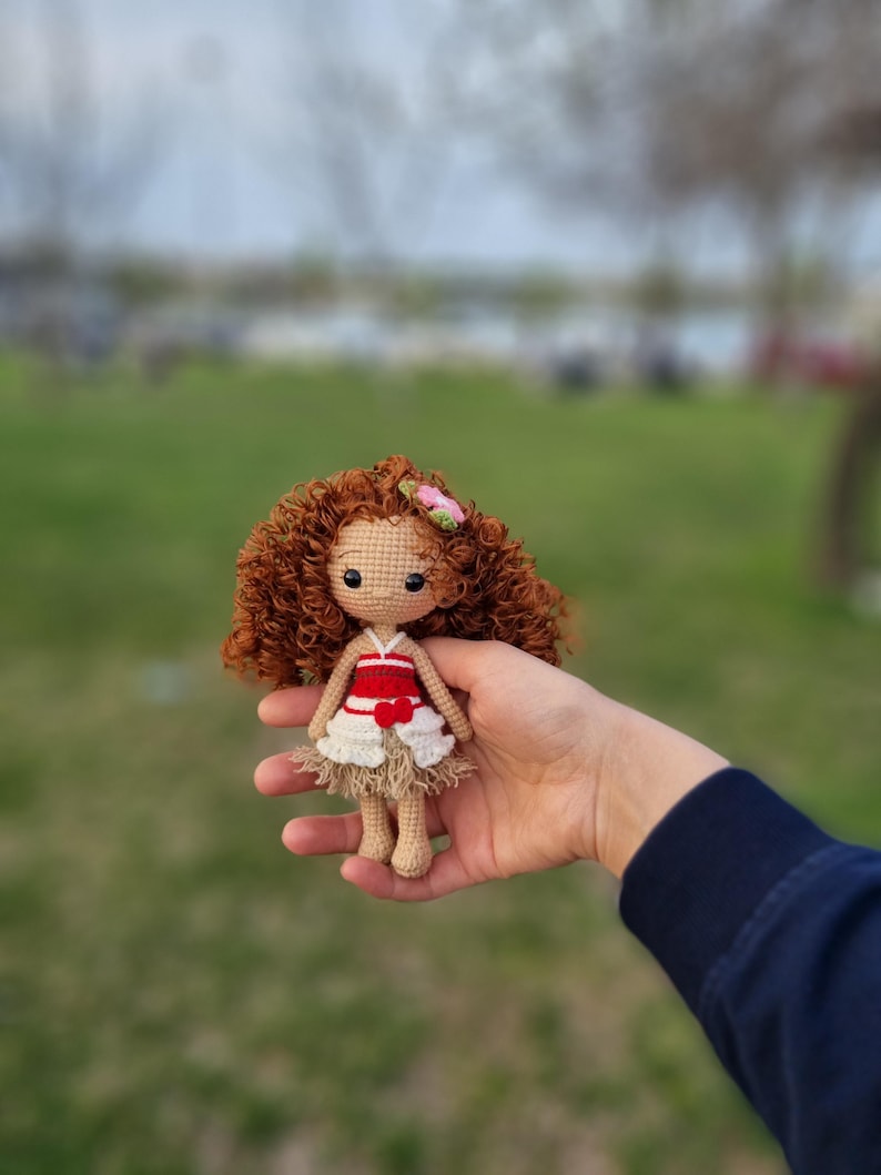 May include: A crocheted doll with brown curly hair, wearing a red and white dress with a flower on her head.