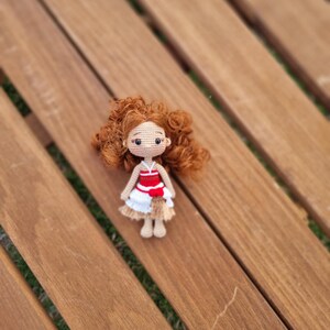 May include: A crocheted doll with brown hair, wearing a red and white dress with a brown skirt. The doll is sitting on a wooden surface.