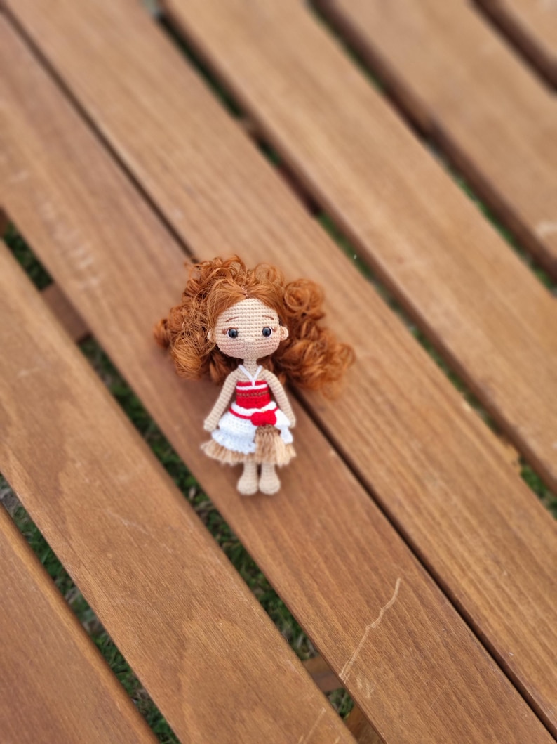 May include: A crocheted doll with brown hair, wearing a red and white dress with a brown skirt. The doll is sitting on a wooden surface.