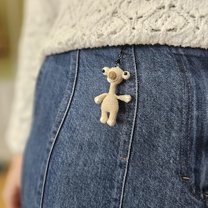 Puede incluir: Un pequeño llavero de extraterrestre tejido en beige con ojos negros y una expresión sonriente. El extraterrestre está sujeto a un anillo de metal negro, colgando de una falda vaquera azul. El fondo incluye un suéter blanco de punto abierto.