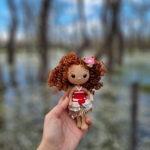 May include: A crocheted doll with brown curly hair, wearing a white dress with a red bodice and a pink flower in her hair.