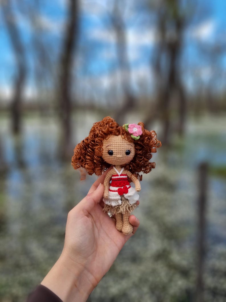 May include: A crocheted doll with brown curly hair, wearing a white dress with a red bodice and a pink flower in her hair.