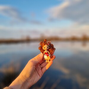 May include: A small crocheted doll with red hair, wearing a red and white dress, is held in a hand against a blurred background of a lake and sky.