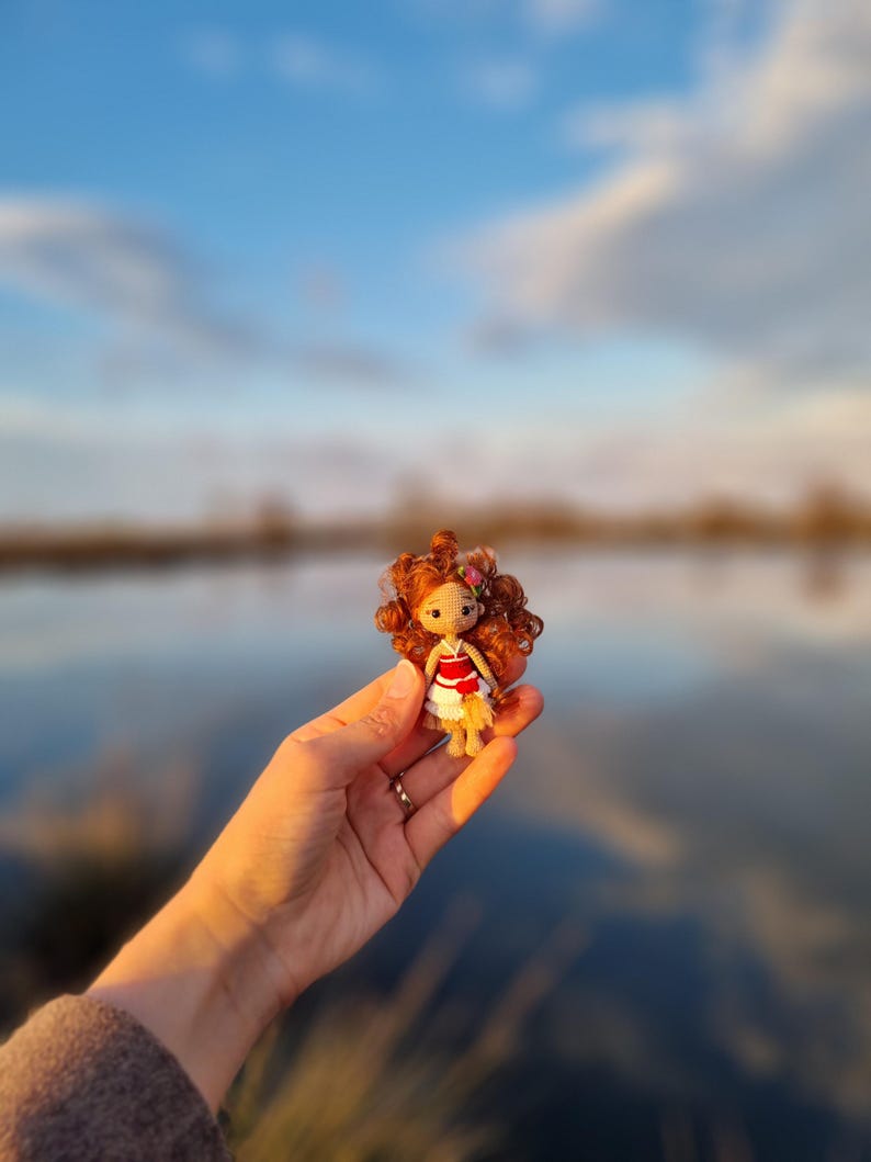 May include: A small crocheted doll with red hair, wearing a red and white dress, is held in a hand against a blurred background of a lake and sky.