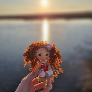 May include: A crocheted doll with brown curly hair, wearing a red and white dress, holding a pink flower. The doll is being held in a hand against a blurry background of a lake and sunset.