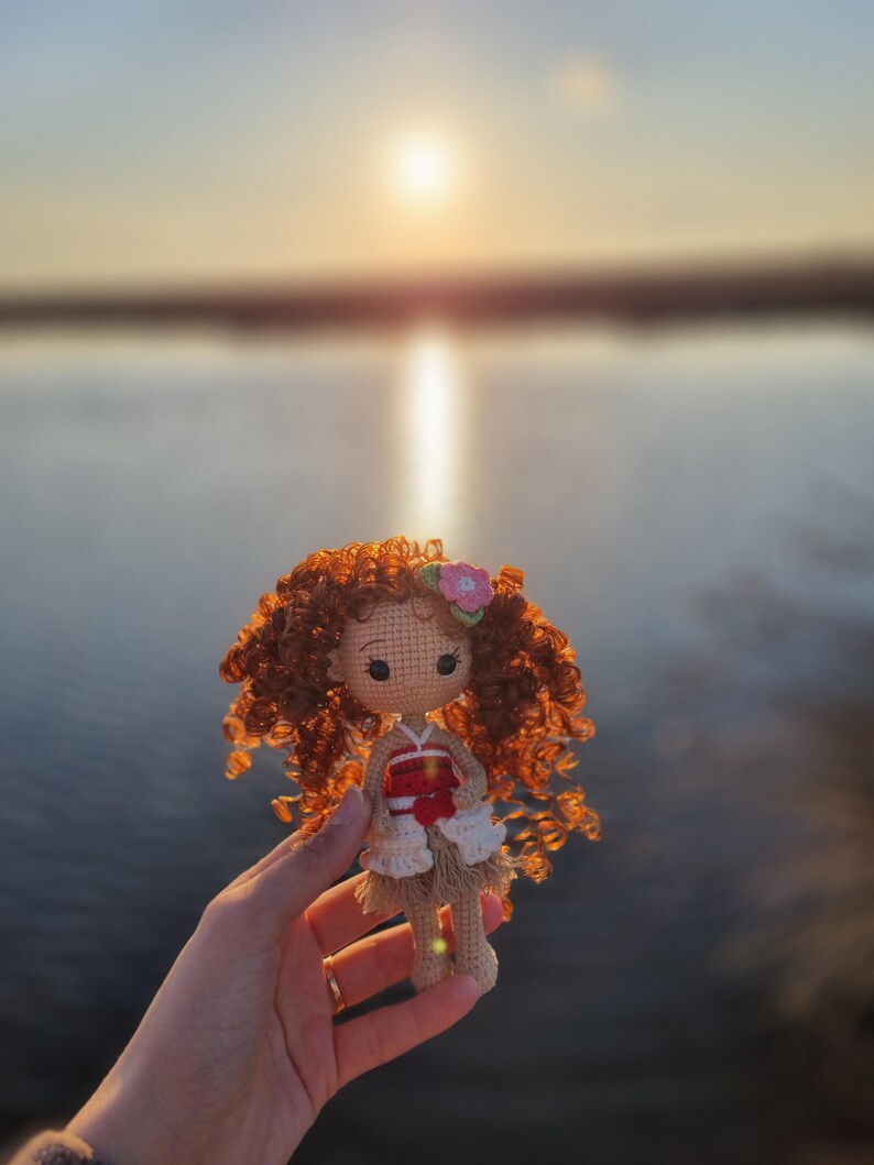 May include: A crocheted doll with brown curly hair, wearing a red and white dress, holding a pink flower. The doll is being held in a hand against a blurry background of a lake and sunset.