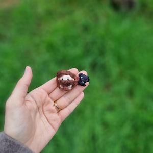 May include: A small brown crocheted figure with a beard and a black crocheted figure with a white face. The figures are being held in a hand.