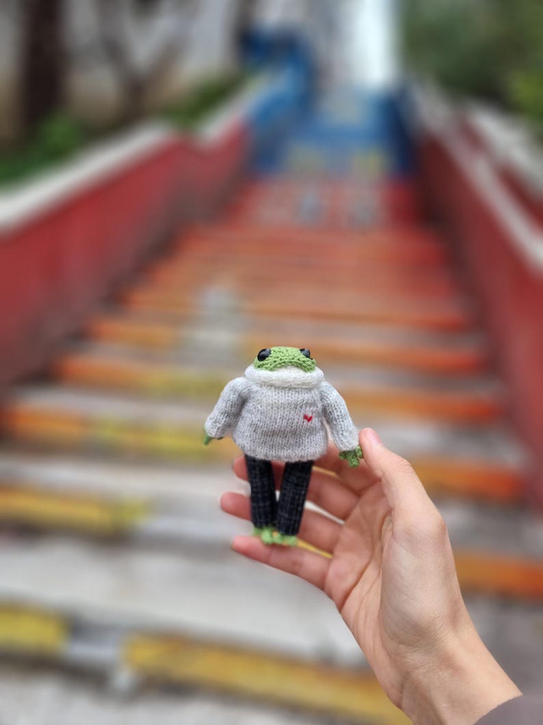 May include: A small green frog wearing a gray sweater with a red heart and black pants. The frog is being held in a person's hand.