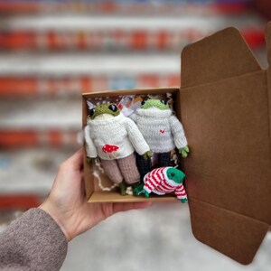 May include: Three knitted frog plushies in a brown cardboard box. The frogs are wearing different colored sweaters. One frog is wearing a white sweater with a red heart, another is wearing a white sweater with a red mushroom, and the third is wearing a red and white striped sweater.
