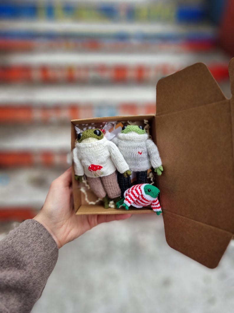 May include: Three knitted frog plushies in a brown cardboard box. The frogs are wearing different colored sweaters. One frog is wearing a white sweater with a red heart, another is wearing a white sweater with a red mushroom, and the third is wearing a red and white striped sweater.