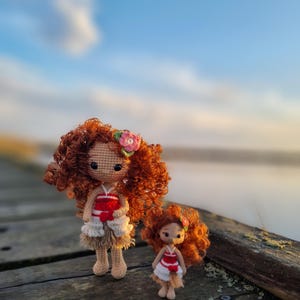 May include: Two crocheted dolls, one larger than the other, are standing on a wooden surface. The larger doll has curly red hair and is wearing a red and white dress with a brown skirt. The smaller doll has curly red hair and is wearing a red and white dress with a brown skirt. The dolls are standing in front of a blurry background of a body of water and a cloudy sky.