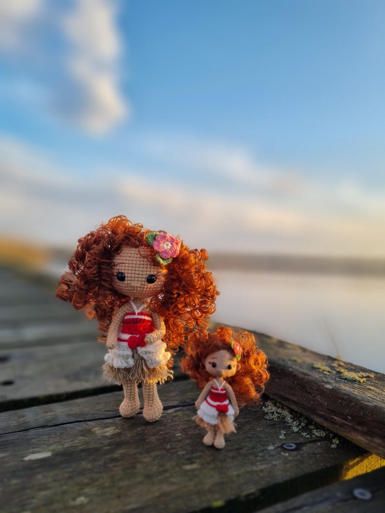 May include: Two crocheted dolls, one larger than the other, are standing on a wooden surface. The larger doll has curly red hair and is wearing a red and white dress with a brown skirt. The smaller doll has curly red hair and is wearing a red and white dress with a brown skirt. The dolls are standing in front of a blurry background of a body of water and a cloudy sky.