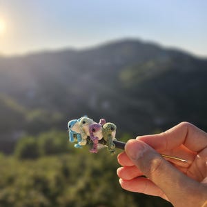 Puede incluir: Una escultura de rana de ganchillo hecha a mano en tonos azules, verdes y rosas. La rana en miniatura se sostiene contra un fondo borroso de una cordillera y un cielo brillante. La rana está unida a una varilla de metal oscuro.