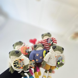 May include: A collection of handmade frog dolls, each with a unique outfit and miniature skateboard. The frogs wear sweaters, hoodies, and shorts in various colors, including blue, white, and red. The skateboards have colorful designs.
