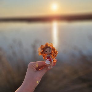 May include: A small crocheted doll with red hair, wearing a red and white dress, is held in a hand against a blurred background of a lake and sunset.
