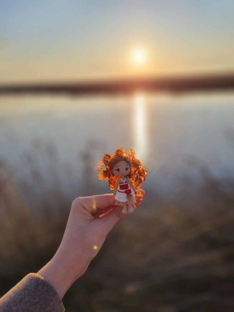 May include: A small crocheted doll with red hair, wearing a red and white dress, is held in a hand against a blurred background of a lake and sunset.