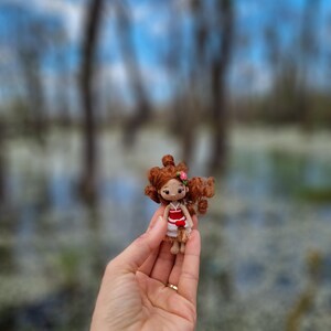 May include: A small crocheted doll with brown hair, a red and white dress, and a pink flower in her hair. The doll is being held in a person's hand.