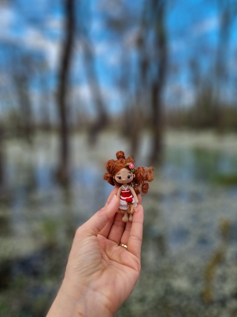 May include: A small crocheted doll with brown hair, a red and white dress, and a pink flower in her hair. The doll is being held in a person's hand.