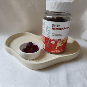 Peut inclure: Une bouteille transparente de gommes LifeCare ImmunXforte+ avec un couvercle blanc, posée sur un plateau beige. Un petit bol blanc contient plusieurs gommes rouge foncé. L'étiquette de la bouteille indique "Avec vitamines, zinc et sélénium".