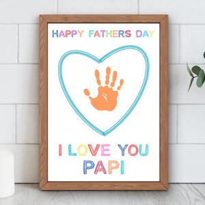 May include: A framed print with a wooden frame. The print features the text "HAPPY FATHERS DAY" in rainbow colours above a blue heart with an orange handprint inside. Below the heart, the text reads "I LOVE YOU PAPI" in colourful letters.