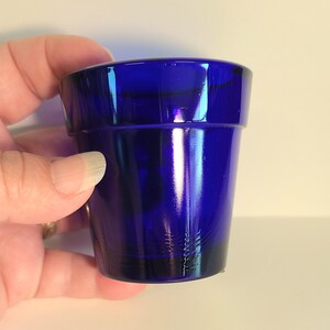 May include: A small, cobalt blue glass planter. The cylindrical pot has a slightly flared rim and a smooth, glossy finish. The dark blue color is consistent throughout the glass, creating a simple, elegant design. The planter is held by a hand.