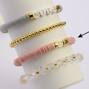 May include: Four bracelets stacked on a white display stand. The top bracelet is gray with white beads that spell "MILA". The second bracelet is gold with small round beads. The third bracelet is pink with white beads. The bottom bracelet is clear with crystal beads and gold accents.