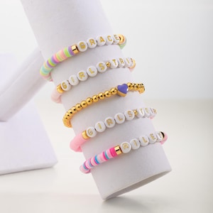 May include: Five colorful beaded bracelets with gold lettering. The bracelets spell out the names "GRACE", "GIRLS TRIP", "GIRLS TRIP", "RILEY". One bracelet has a purple heart charm.