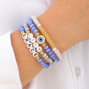 May include: A set of four bracelets featuring blue and white beads, a gold charm with a blue evil eye, and white beads with the names "GRACE", "LILLY" spelled out.