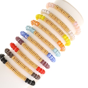 May include: A collection of beaded bracelets in various colors, including black, red, purple, blue, light blue, orange, yellow, and pink. Each bracelet features a gold-colored central band. The bracelets are displayed on a white cylindrical surface.