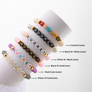 May include: Six colourful beaded bracelets with the name "Audrey" spelled out in different coloured beads. The bracelets are stacked on top of each other, with the top bracelet having colourful beads, the second bracelet having black beads with white letters, the third bracelet having white beads with black letters, the fourth bracelet having black beads with gold letters, the fifth bracelet having multi-coloured beads, and the bottom bracelet having white beads with gold letters.