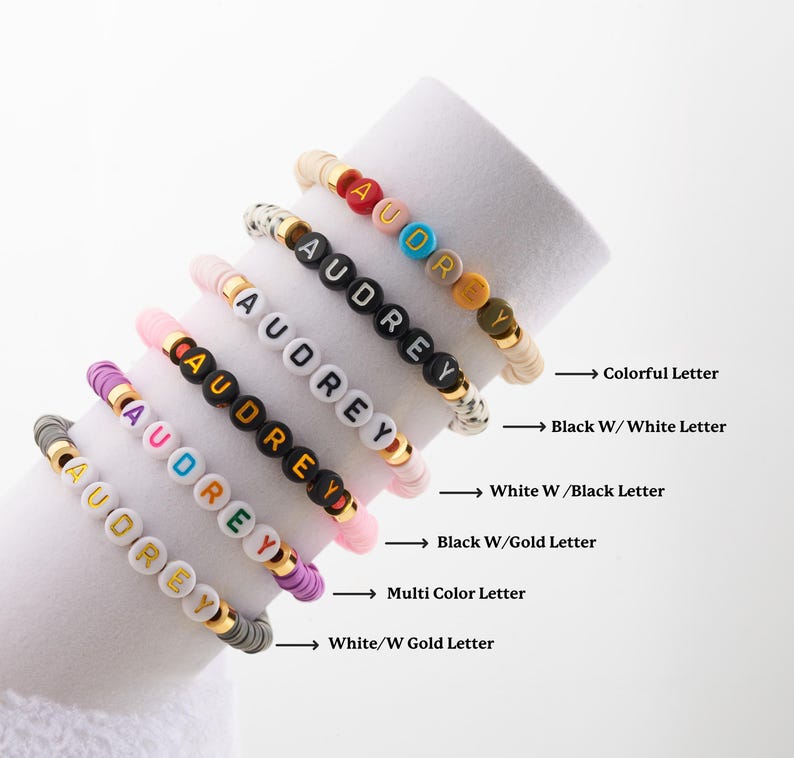 May include: Six colourful beaded bracelets with the name "Audrey" spelled out in different coloured beads. The bracelets are stacked on top of each other, with the top bracelet having colourful beads, the second bracelet having black beads with white letters, the third bracelet having white beads with black letters, the fourth bracelet having black beads with gold letters, the fifth bracelet having multi-coloured beads, and the bottom bracelet having white beads with gold letters.