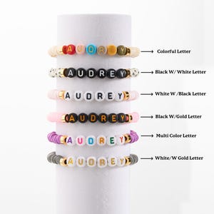 May include: Six stacked bracelets with the name "AUDREY" spelled out in colourful beads. The bracelets are made with different coloured beads and letter colours, including black, white, gold, and multi-coloured.